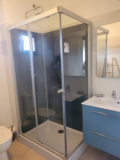 Salle de bain moderne avec douche vitrée, meuble vasque bleu, toilettes et miroir à l'appartement Au Golfe de lava en Corse