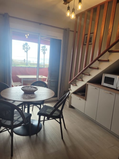 Salle à manger d'un appartement moderne en Corse avec table ronde, balcon vue mer, escalier en bois et palmiers visibles dehors