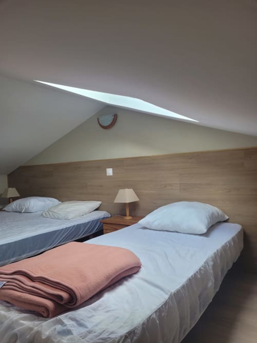Chambre mansardée dans un gîte corse avec deux lits, tête de lit en bois, couverture rose et lucarne