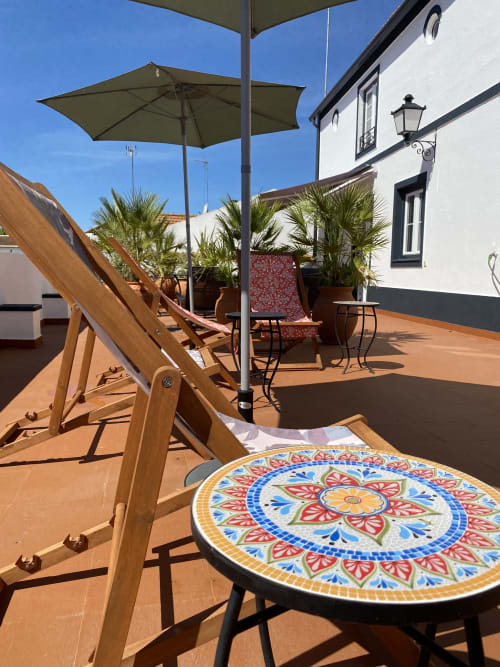 Terraço ensolarado do Patio Da Villa com espreguiçadeiras de madeira, mesa decorativa em azulejo, guarda-sol, plantas de palmeira e branco