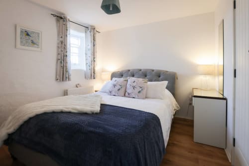 Bright bedroom at Ballymultimber Cottages with gray headboard bed, white linens, patterned pillows, wooden floor, and window