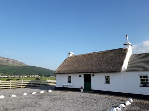 Traditional white-washed cottage with thatched roof at Ballymultimber Cottages, Limavady, set against green mountains.