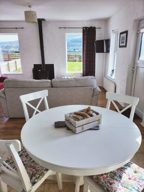 Open-plan dining and sitting area at Ballymultimber Cottages with round white table, grey sofa, wood stove, and mountain