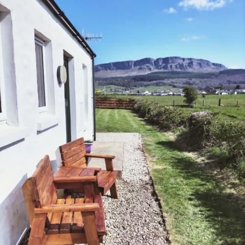 White cottage with wooden chairs on gravel patio overlooking green fields and mountains at Ballymultimber Cottages, Limavady