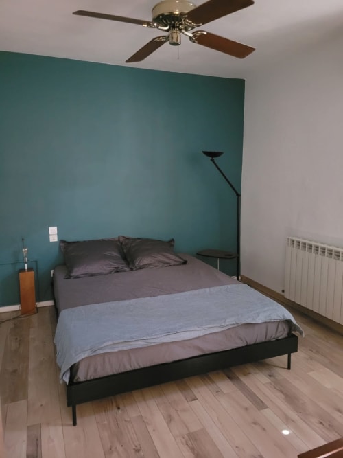 Modern bedroom in La Conciergerie De Franck villa with turquoise accent wall, queen bed, ceiling fan, and wooden flooring