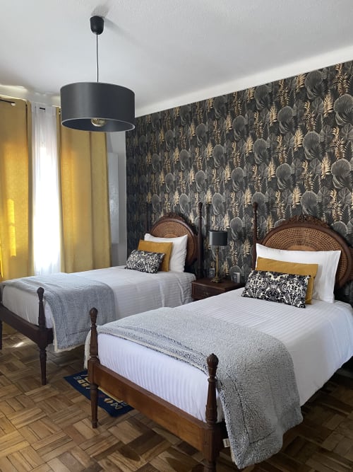Twin bedroom with wooden beds, rattan headboards, gold and black patterned wallpaper, yellow curtains, and pendant light at