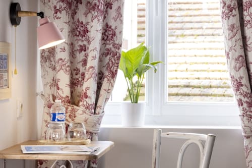 Chambre d'hôtel lumineuse à l'Hôtel De Lion Sur Mer avec rideaux à fleurs, lampe de bureau, plante en pot et vue sur Lion sur Mer