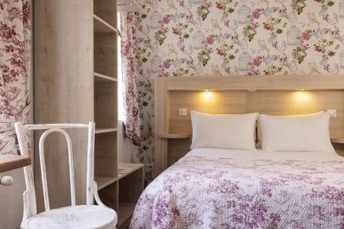 Chambre double standard à l'Hotel De Lion Sur Mer avec papier peint fleuri, chaise blanche, lit en bois avec literie matelassée rose