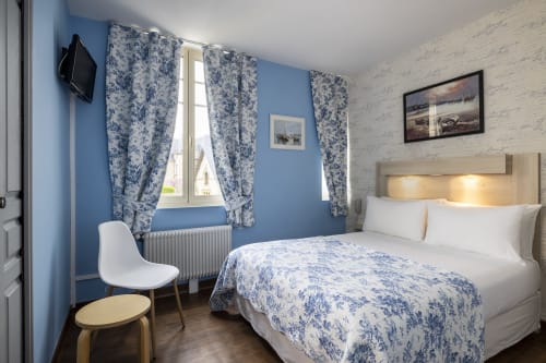Chambre double confort à l'Hôtel De Lion Sur Mer avec murs bleus, rideaux à motifs bleus, literie blanche et mur d'accent en bois