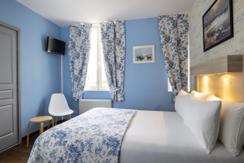 Chambre double confortable à l'Hôtel De Lion Sur Mer avec murs bleus, literie blanche, rideaux à motifs et télévision écran plat
