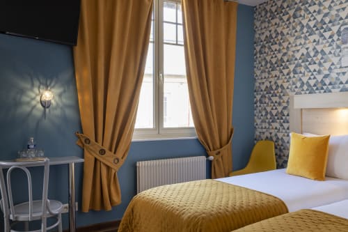 Chambre triple à l'Hôtel De Lion Sur Mer aux murs bleus, rideaux dorés, accents jaunes et vue sur la ville par de grandes fenêtres
