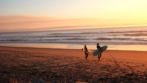 Two surfers stood on a sandy beach about to enter the sea.