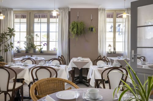 Élégante salle à manger de l'Hôtel De Lion Sur Mer avec nappes blanches, chaises bistrot, grandes fenêtres et plantes suspendues
