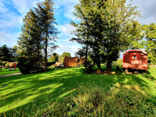 Red vintage caravan and wooden lodge surrounded by green fields and tall trees at Les Refuges Du Chalet in Jalhay, Belgium.