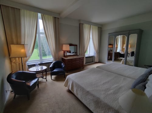 Chambre élégante du Château d'Hassonville avec vue sur jardin, lits jumeaux, fauteuils en cuir noir et grandes fenêtres