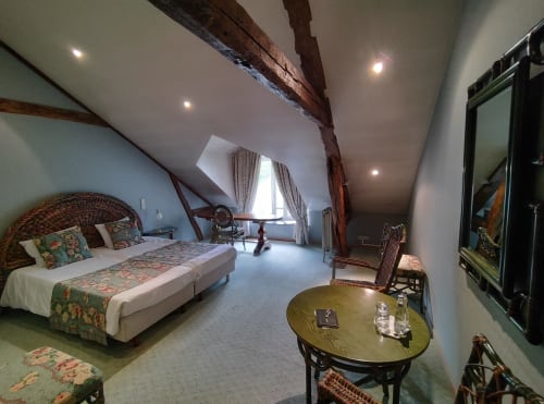 Spacieuse chambre mansardée au Château d'Hassonville avec poutres apparentes, tête de lit en rotin et mobilier élégant