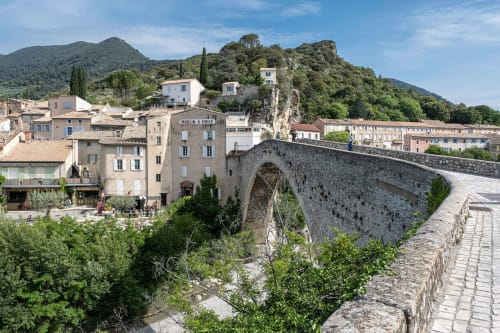 Medieval stone bridge spanning valley in Grignan, France with terracotta-roofed village buildings and green hills
