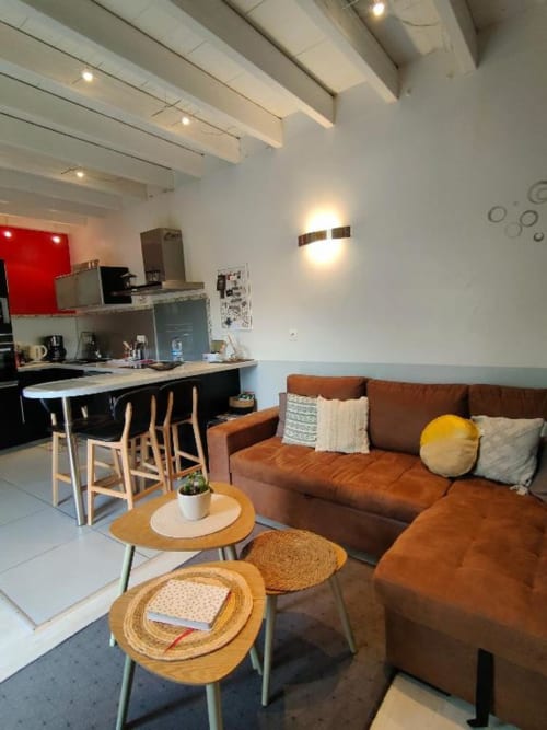 Séjour moderne ouvert dans la villa de Dinan avec canapé marron, tables d'appoint en bois, bar cuisine avec sièges et poutres apparentes