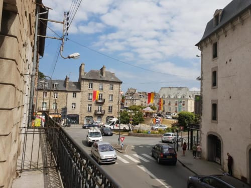Place médiévale à Dinan avec bâtiments en pierre, bannières colorées, voitures garées et vue animée de la rue