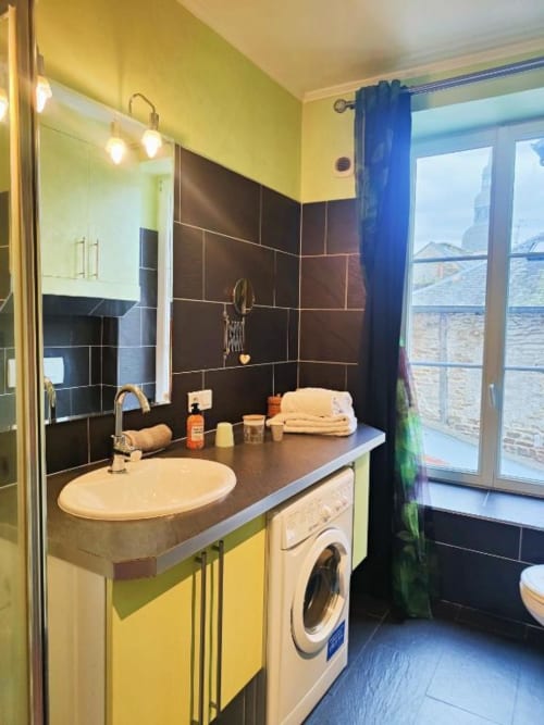 Salle de bains moderne aux murs jaunes, carrelage noir, lavabo rond, machine à laver et vue sur balcon à la villa de Dinan