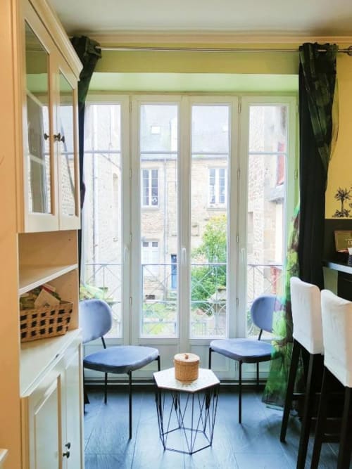 Studio lumineux à Dinan avec vue sur la ville, chaises bleues, petite table et portes-fenêtres donnant sur les bâtiments historiques
