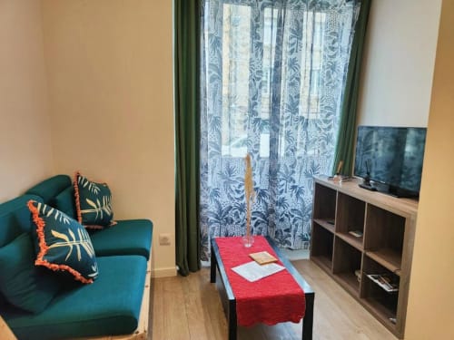 Studio cosy à Dinan avec canapé bleu-vert, télévision et balcon avec vue sur la ville et rideaux en dentelle
