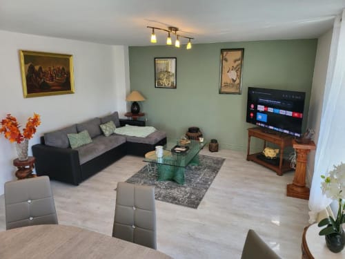 Salon moderne de l'appartement de Dinan avec canapé gris, mur d'accent vert, smart TV et table basse en verre