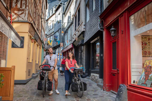 Couple à vélo dans la charmante rue de Honfleur près de la maison d'hôtes Les Maisons de Pierre et Valérie, bâtiments colorés et portes rouges