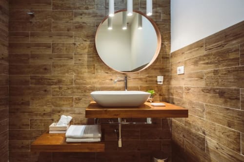 Moderno bagno di Bluemoon Sardegna Cagliari con piastrelle effetto legno, lavabo bianco a coppa e specchio rotondo con lampada sospesa