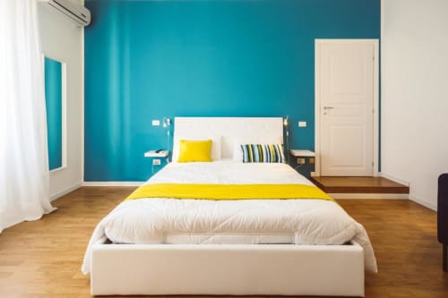 Camera da letto moderna a Bluemoon Sardegna con parete azzurra, letto bianco, dettagli gialli, pavimenti in legno e vista città