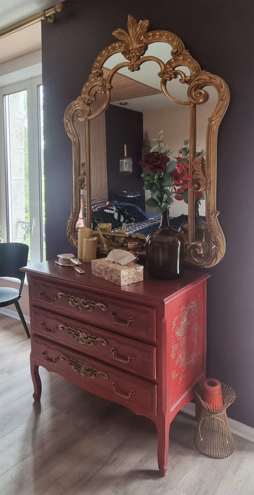 Miroir doré aux ornements raffinés au-dessus d'une commode rouge avec fleurs, dans une suite de Les Forges Du Lou à Jugon-les-Lacs