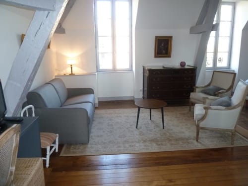 Dormitorio abuhardillado con vigas vistas, sofá gris, cómoda de madera y grandes ventanales en Château De Clusors, ST MENOUX