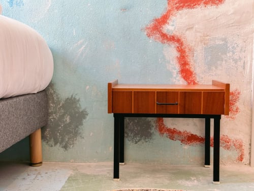 Mesita de noche de mediados de siglo en madera con patas negras contra pared pastel desgastada en Hotel La Prison, Béziers