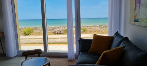 Coastal cottage interior with sea view through glass doors, dark sofa, and sandy beach at RELAIS DES ILES, Saint-Marcouf