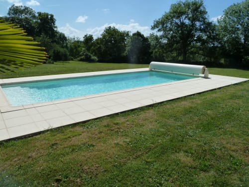 Piscina rectangular turquesa con terraza blanca rodeada de césped y árboles en el Château De Clusors, campiña francesa