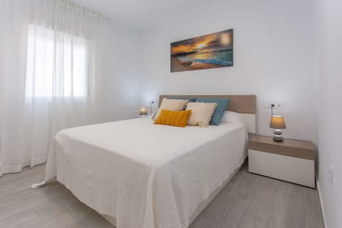 Dormitorio luminoso en apartamento Wehost Costaluz con cama blanca, cabecero de madera, obra de arte con atardecer oceánico y luz natural