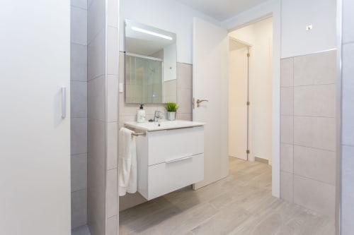 Cuarto de baño moderno en apartamento Wehost Costaluz con vanidad blanca, espejo, ducha y piso de madera clara en Puerto Santa María