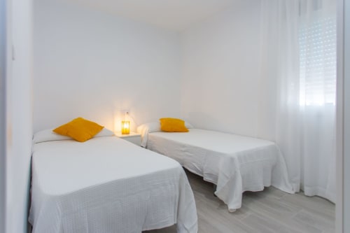 Luminosa habitación doble en Wehost Costaluz con sábanas blancas, almohadas doradas e iluminación ambiental en Puerto Santa María