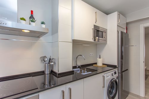 Cocina moderna blanca con encimeras negras, electrodomésticos de acero inoxidable y lavadora en apartamento de Puerto Santa María