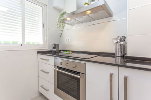 Cocina moderna blanca con horno de acero inoxidable, encimera negra y plantas colgantes en el apartamento Las Dunas2 de Wehost