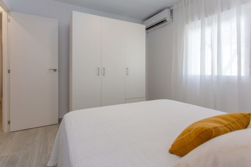 Dormitorio moderno y luminoso con cama blanca, almohada naranja, amplio armario, aire acondicionado y cortinas translúcidas en apartamento Las Dunas,
