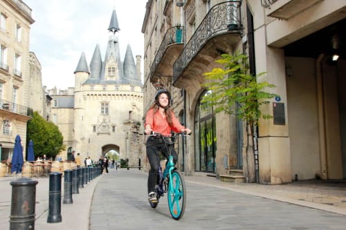 Femme à vélo sur un vélo turquoise traversant une rue médiévale de Bordeaux devant une tour de château, secteur Le Prieuré Du Preux