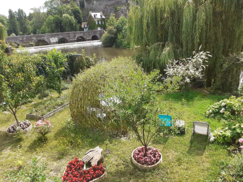 Le jardin du refuge des Alpes mancelles surplombant la Sarthe avec fleurs, saule pleureur et pont de pierre
