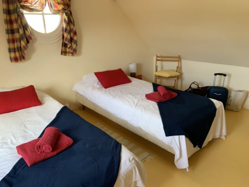 Zweibettzimmer mit roten Kissen, dunkelblauem Bettwäsche, gerollten roten Handtüchern, Holzstuhl und karierten Vorhängen im La Croix De La