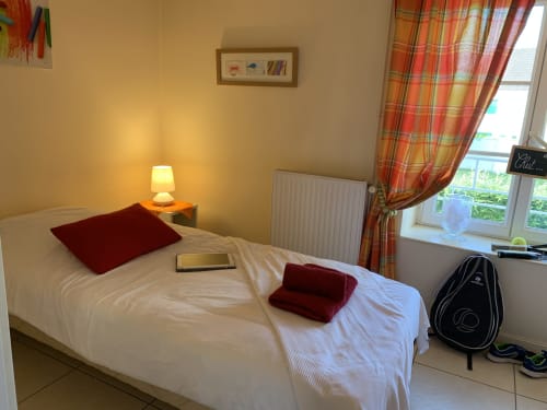 Gemütliches Schlafzimmer in La Croix De La Jarrie mit weißem Bett, burgunderfarbenen Kissen, Schreibtischlampe und karierten Vorhängen mit Gartenblick