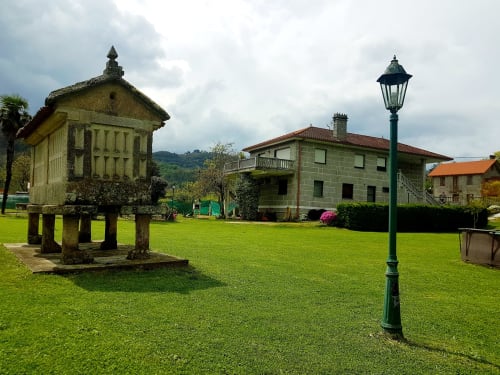 Jardín de la casa de alquiler vacacional O Eido de Celia, en Guláns, Ponteareas, con un hórreo tradicional gallego, farola antigua y césped bien cuidado, junto a una casa de piedra rural en un entorno verde y tranquilo.