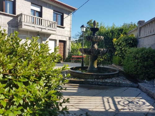 Patio de la casa de alquiler vacacional O Eido de Celia, en Guláns, Ponteareas, (Galicia) con una fuente de piedra de varios niveles rodeada de vegetación, y una casa de piedra rural al fondo, en un día soleado.