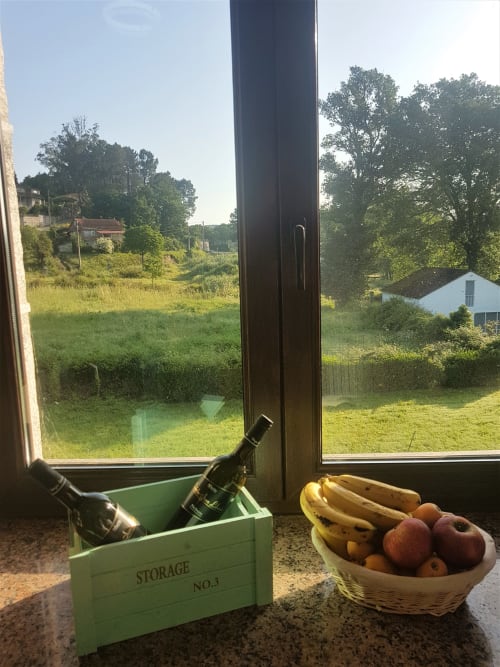 Vista desde una ventana de la casa de alquiler vacacional O Eido de Celia, en Guláns, Ponteareas, Galicia, con una caja de vino y una cesta de frutas en el alféizar, y un paisaje rural con árboles y una casa de campo al fondo.