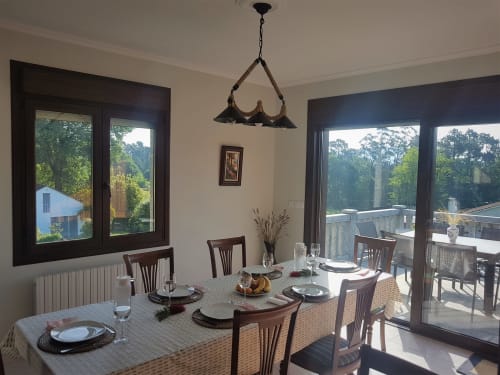 Comedor interior de la casa de alquiler vacacional O Eido de Celia, en Guláns, Ponteareas, Galicia, con mesa preparada para una comida y grandes ventanales que ofrecen vistas al exterior y a la terraza.