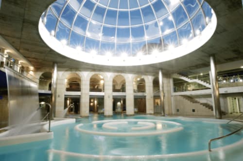 Interior del Palacio del Agua en Mondariz Balneario, con una gran piscina termal bajo una cúpula de vidrio iluminada, rodeada de columnas y arcos, ideal para disfrutar de aguas termales en un entorno relajante y lujoso.